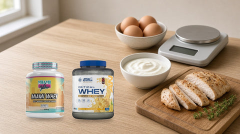 Proteine din zer Miami Whey și Applied Nutrition Critical Whey pe un blat de bucătărie, lângă un cântar de bucătărie, un bol cu ouă, un bol cu iaurt și felii de piept de pui gătit