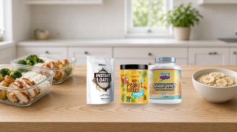 Ovăz instant BiotechUSA, unt de arahide OstroVit și proteină Miami Whey pe un blat de bucătărie, între caserole cu pui, orez, broccoli și cartofi și un bol cu ovăz și banană